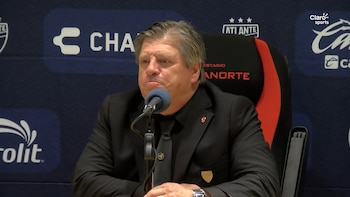 El director técnico Miguel Herrera, vestido con traje y corbata negros, permanece sentado frente a un micrófono