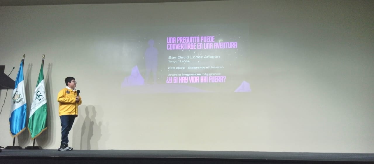 David López Aragón, de 11 años, presenta su ponencia sobre la exploración del universo junto a las banderas institucionales en un escenario. (Cortesía: Pamela López)
