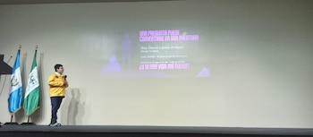 Un niño con chaqueta amarilla habla con un micrófono ante una pantalla proyectada y dos banderas de Guatemala en un escenario
