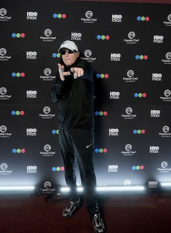 Hombre con gorra blanca, gafas de sol y ropa deportiva negra apunta hacia adelante ante un fondo con logotipos de MasterChef Celebrity y HBO Max