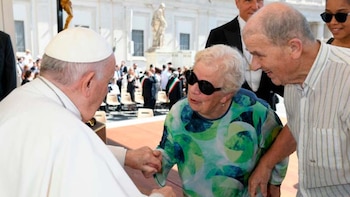 Primer plano del Papa Francisco, un hombre mayor, sentado y vestido con una sotana blanca, con la mano derecha levantada hacia la oreja, y un micrófono delante. Se ve una cruz pectoral