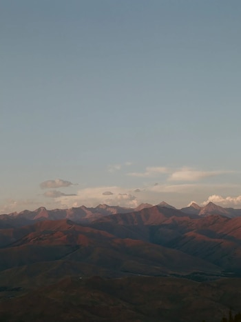 Una extensa cordillera con cumbres y laderas de tonos rojizos y ocres. Arriba, un cielo azul claro con nubes cúmulos blancas