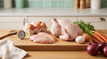 Un pollo entero crudo, dos pechugas, huevos, zanahorias, cebollas moradas, un termómetro de cocina, una cuchara de madera y un paño sobre una tabla.