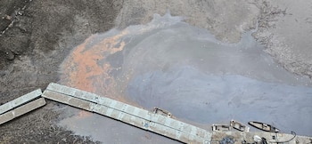 Superficie de agua con manchas de color gris iridiscente y rojizo, estructuras rectangulares de concreto, y dos botes volcados con residuos en primer plano