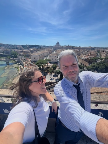 Un hombre y una mujer posan en una selfie con la Basílica de San Pedro, edificios históricos, un río y puentes bajo un cielo azul claro