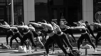 Fotografía en blanco y negro de un grupo de mujeres descalzas en ropa deportiva oscura realizando una pose de yoga al aire libre sobre esterillas.