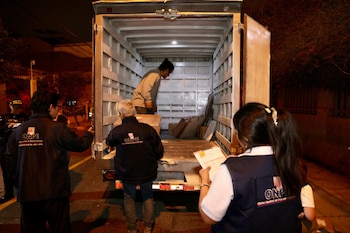 Varias personas, algunas con chalecos de ONPE, cargan cajas en la parte trasera abierta de un camión blanco, en una calle oscura por la noche