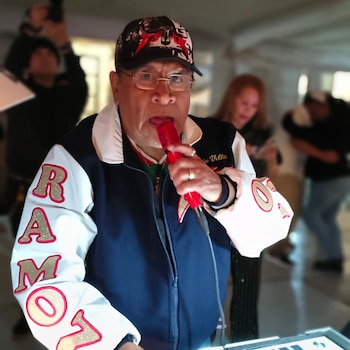 Un hombre con gafas, gorra y chaqueta deportiva sostiene un micrófono rojo cerca de su boca frente a una consola de audio