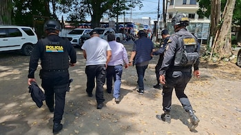 Dos agentes uniformados de la Policía Nacional Civil caminan junto a tres hombres vestidos de civil por un camino de tierra