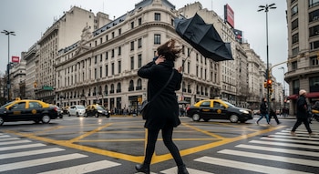 Vista trasera de una persona con abrigo oscuro cruzando una calle en Buenos Aires, sujetando un paraguas negro invertido por el viento. Taxis y edificios al fondo.