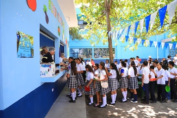 Estudiantes en uniforme hacen fila en un puesto de comida escolar azul con decoraciones de vegetales, siendo atendidos por personal. Hay banderines colgando