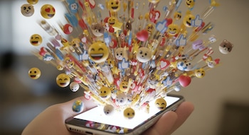 Una mano sostiene un smartphone del cual emergen numerosos emojis de diversos colores y formas, creando un efecto de explosión visual sobre un fondo borroso.
