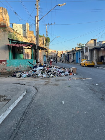 Una calle con una gran pila de basura en el centro, flanqueada por edificios antiguos y deteriorados, con un coche amarillo aparcado