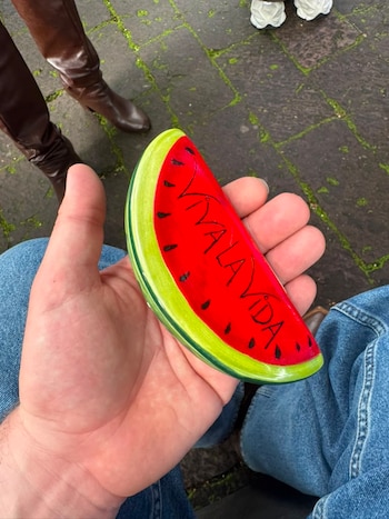 Mano sujeta objeto decorativo con forma de sandía roja, semillas negras, borde verde y texto "VIVA LA VIDA". La persona viste jeans. Fondo de baldosas y botas