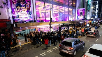 Multitud de personas hace fila frente a un edificio iluminado con carteles de 'Charlie y la Fábrica de Chocolate' y 'Tabaris' en una calle urbana de noche con coches.