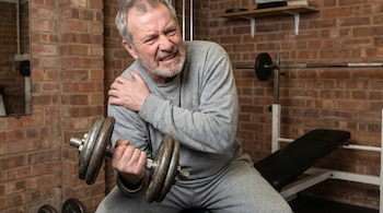 Un hombre de barba y cabello gris, vistiendo ropa deportiva gris, levanta una mancuerna mientras se agarra el hombro con expresión de dolor.
