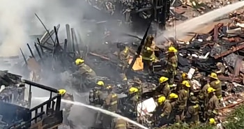 Bomberos con cascos amarillos y uniformes oscuros rocían agua sobre los restos humeantes y carbonizados de una estructura completamente quemada en un incendio