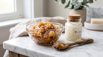 Un cuenco de vidrio con exfoliante de azúcar, una cuchara de madera y un frasco de aceite de coco sobre una mesa de mármol.