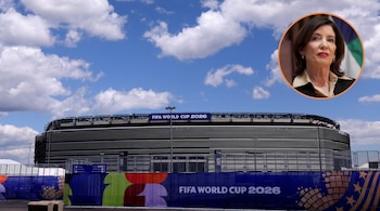 Vista exterior de un estadio moderno bajo un cielo azul con nubes, con banners de "FIFA World Cup 2026". En la esquina superior derecha, retrato de Kathy Hochul.