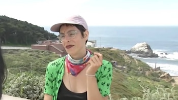 Una persona joven con gafas y gorra rosa, vistiendo una camisa verde y pañuelo, de pie frente a un paisaje costero con el océano y formaciones rocosas