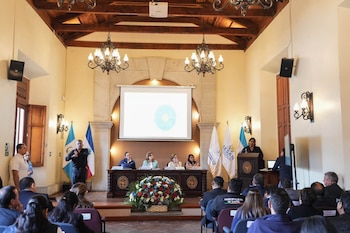 Personas reunidas en un salón con una mesa directiva, un orador en un podio, un intérprete de lengua de señas, banderas y una pantalla