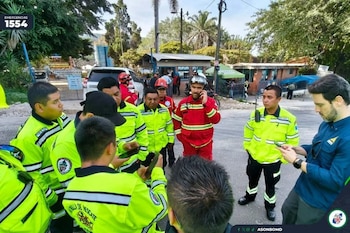 Grupo de personas con uniformes de emergencia amarillos, rojos y azules en una calle. Un hombre habla por teléfono, otros revisan sus móviles. Hay árboles y edificios
