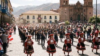 Personas desfilan en una calle empedrada, algunas vestidas con uniformes y trajes tradicionales, otras observan; se ven banderas peruanas, edificios coloniales y montañas.