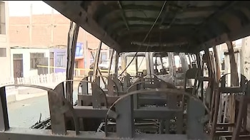 Interior de un autobús calcinado, asientos y estructura metálicos quemados, con ventanas rotas. Al fondo, se observan edificios y un vehículo