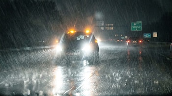Vista frontal de un auto con faros y balizas encendidas, avanzando en una carretera bajo una lluvia torrencial. Las gotas de agua son muy visibles.