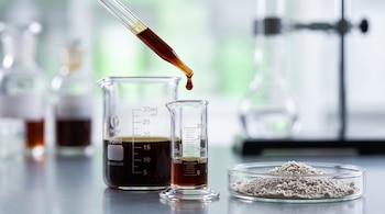 Un gotero con líquido ámbar sobre un cilindro graduado, un vaso de precipitados y una placa de Petri con polvo blanco en un laboratorio.