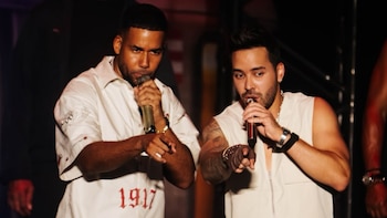 Romeo Santos y Prince Royce sostienen micrófonos y señalan hacia adelante vestidos con ropa blanca sobre un escenario