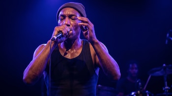 Un hombre con gorro de lana y camiseta sin mangas canta con los ojos cerrados frente a un micrófono con iluminación azul.