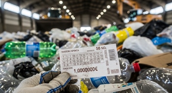 Una mano enguantada sostiene un billete de lotería que muestra números y un millón de euros. Al fondo, una pila de basura con botellas de plástico.