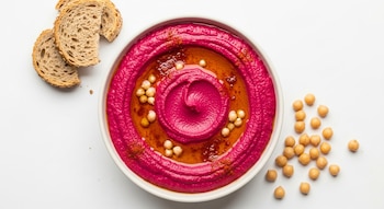 Un bowl de hummus de remolacha rosa brillante, adornado con aceite, garbanzos y pimentón. Rebanadas de pan integral y garbanzos sueltos están al lado sobre fondo blanco.