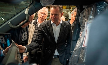 Primer plano de un hombre con traje oscuro y camisa blanca que toca el interior de un vehículo. Otros hombres se ven detrás. Parte del vehículo está cubierta con plástico