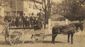 Fotografía sepia. Un grupo de hombres blancos, algunos con rifles, está sobre un carro de madera tirado por un caballo, con un edificio de fondo