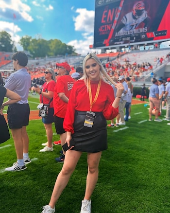 La periodista Dianna Russini sonríe en el campo de un estadio de fútbol americano, llevando blusa roja y falda negra. Al fondo, gradas y un marcador