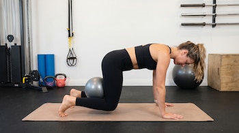 Una mujer con ropa deportiva negra realiza un ejercicio en cuadrupedia sobre una colchoneta, con una pequeña pelota plateada entre sus rodillas ligeramente elevadas. El fondo es un gimnasio con equipo.