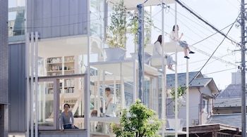 Casa de múltiples niveles con paredes de cristal y estructura metálica, personas, estanterías con libros, plantas en macetas, postes y cables eléctricos en exterior.