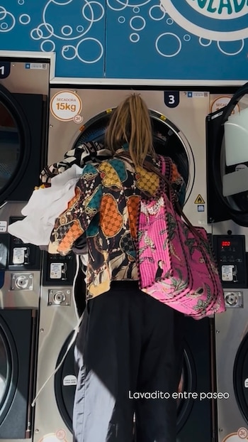Una mujer de espalda introduce ropa en una secadora dentro de una lavandería pública mientras lleva un bolso grande de color rosa