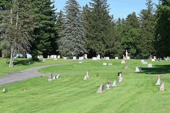 Cementerio con lápidas de piedra en un campo de césped verde. Un camino serpentea. Árboles coníferos altos forman un denso fondo bajo un cielo azul