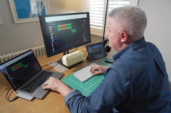 Hombre con pelo gris en camisa de mezclilla trabajando en un escritorio con portátil, monitor, tablet, teclado y altavoz, produciendo música