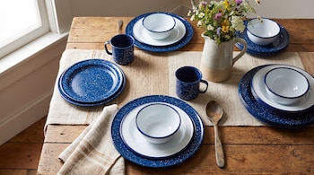 Platos, cuencos y tazas de peltre azul con motas sobre un camino de mesa junto a una cuchara de madera y un jarrón con flores.