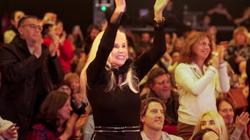 Susana Giménez, con cabello rubio y top negro, con los brazos levantados y las manos abiertas aplaudiendo, sonriendo ampliamente entre una multitud en un teatro