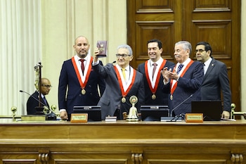 Seis hombres en trajes formales con bandas bicolores y medallas en un recinto. El hombre central alza un libro. Hay micrófonos, campana y crucifijo