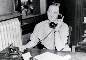 Fotografía en blanco y negro de una mujer sentada junto a un escritorio hablando por un teléfono antiguo con papeles frente a ella