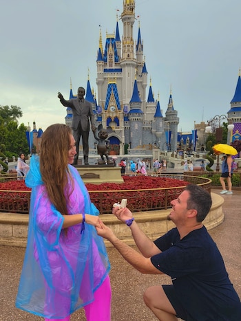 Un hombre arrodillado le ofrece un anillo a una mujer con impermeable azul, con un castillo y una estatua de fondo