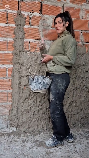 Mujer de pie frente a pared de ladrillo con mezcla de cemento. Sostiene un balde de material y levanta el pulgar. Viste ropa de trabajo manchada