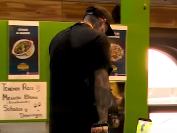 Hombre de espaldas con playera negra y gorra, mostrando tatuajes en el brazo, en un interior con paredes verdes, carteles de comida y dos teléfonos móviles abajo