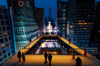 Vista elevada de Times Square al anochecer. Personas en una plataforma observan la calle, tráfico y pantallas. Rascacielos iluminados, One Times Square con reloj